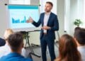 In the well-lit conference room of a modern office, a professional businessman stands confidently as he delivers a compelling presentation. He engages directly with his audience, pointing towards the colorful bar graphs on the screen that likely represent the company's financial growth or market analysis. The audience, comprising a group of attentive listeners in business attire, is focused on the speaker, anticipating valuable insights. The dynamic atmosphere suggests a collaborative corporate environment where ideas and strategies are actively discussed.