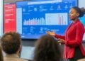 In a well-lit corporate meeting room, a professional woman stands confidently as she presents complex statistical data displayed on a large screen behind her. Her audience, a group of attentive colleagues, listens as she explains the significance of the data trends and graphs, suggestive of a strategic business review or financial update session. Her poised demeanor and the engagement of her audience illustrate a crucial business discourse aimed at driving company performance.