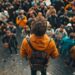 In this dynamic photo, we see a solitary individual dressed in a bright yellow jacket and backpack standing above a sea of people. Positioned in a unique vantage point, the person appears as if he's addressing or leading the crowd, which is composed of various individuals focused in different directions. This image captures a moment of leadership or prominence, amidst an event that draws together a diverse group of spectators.