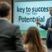 In a well-lit conference room, a confident male businessman engages with a group of attentive professionals during a motivational seminar. He stands in front of a large digital screen displaying the text, 'key to success, Your Your Potential,' gesturing with his hand to emphasize his points. This inspiring scene captures the essence of professional development and leadership in a corporate setting, where ideas are shared and potential is unlocked.