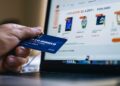 In this photo, we see a person's hand holding a credit card in front of a laptop displaying an online store's webpage. The image captures the moment of an e-commerce transaction, signifying the convenience and simplicity of purchasing goods and services in the digital age. This common scene highlights the blend of technology and commerce and how it has transformed the way consumers engage with retailers.
