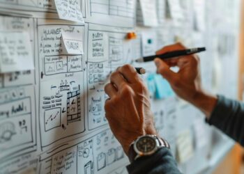 In a scene of focused productivity, an individual is captured in the act of planning and strategizing. With a marker in hand and a wall filled with meticulously arranged sticky notes and flowcharts, they contemplate the next step in their elaborate scheme. The attentive gaze suggests a mind deeply engaged in problem solving, outlining a path to success on the whiteboard that serves as a canvas to their thoughts.