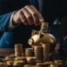 In a dimly lit office, a professional businessman dressed in a dark suit carefully places coins into a shining golden piggy bank, positioned beside a stack of similarly gleaming coins. Behind him, a laptop displays a graph with upward trends, emphasizing the concept of growth and success in financial management. This act represents prudent and calculated financial planning, where even the smallest additions contribute to larger savings and investment strategies.
