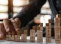 In this photo, a person's hand is depicted placing wooden blocks in a ascending pattern on a table, which artistically represents growth and progression. The warm, sunlit setting creates a focused and dynamic atmosphere, hinting at a strategic business planning session. This visual metaphor elegantly symbolizes the building blocks of success, highlighting the importance of careful planning and incremental advancement in achieving long-term goals.