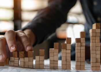 In this photo, a person's hand is depicted placing wooden blocks in a ascending pattern on a table, which artistically represents growth and progression. The warm, sunlit setting creates a focused and dynamic atmosphere, hinting at a strategic business planning session. This visual metaphor elegantly symbolizes the building blocks of success, highlighting the importance of careful planning and incremental advancement in achieving long-term goals.