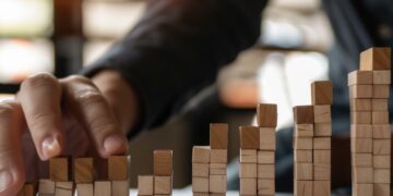 In this photo, a person's hand is depicted placing wooden blocks in a ascending pattern on a table, which artistically represents growth and progression. The warm, sunlit setting creates a focused and dynamic atmosphere, hinting at a strategic business planning session. This visual metaphor elegantly symbolizes the building blocks of success, highlighting the importance of careful planning and incremental advancement in achieving long-term goals.