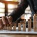 In this photo, a person's hand is depicted placing wooden blocks in a ascending pattern on a table, which artistically represents growth and progression. The warm, sunlit setting creates a focused and dynamic atmosphere, hinting at a strategic business planning session. This visual metaphor elegantly symbolizes the building blocks of success, highlighting the importance of careful planning and incremental advancement in achieving long-term goals.