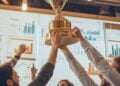 In a lively office setting, a group of enthusiastic colleagues raises a large championship trophy aloft, celebrating a significant achievement. Their expressions of joy and success shine brightly as they stand beneath office lights, surrounded by charts and data that underscore their hard work. This victorious moment reflects their teamwork and dedication to their goals, fostering a spirit of camaraderie and achievement within the workplace.