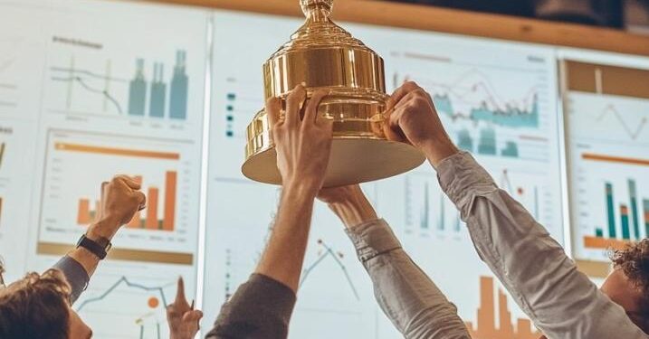 In a lively office setting, a group of enthusiastic colleagues raises a large championship trophy aloft, celebrating a significant achievement. Their expressions of joy and success shine brightly as they stand beneath office lights, surrounded by charts and data that underscore their hard work. This victorious moment reflects their teamwork and dedication to their goals, fostering a spirit of camaraderie and achievement within the workplace.