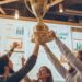 In a lively office setting, a group of enthusiastic colleagues raises a large championship trophy aloft, celebrating a significant achievement. Their expressions of joy and success shine brightly as they stand beneath office lights, surrounded by charts and data that underscore their hard work. This victorious moment reflects their teamwork and dedication to their goals, fostering a spirit of camaraderie and achievement within the workplace.